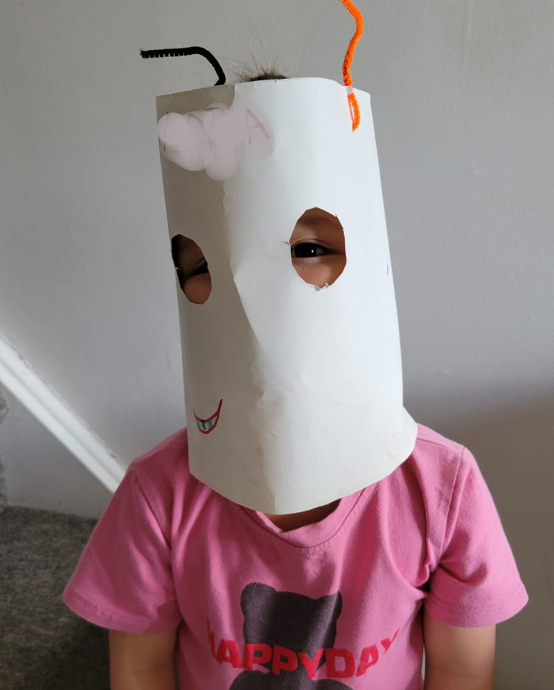 Image shows Baobao wearing a white mask that fully covers her face. It is made of white construction paper and has two large eye-holes, and two antennae made of pipe-cleaners, one white and one black.